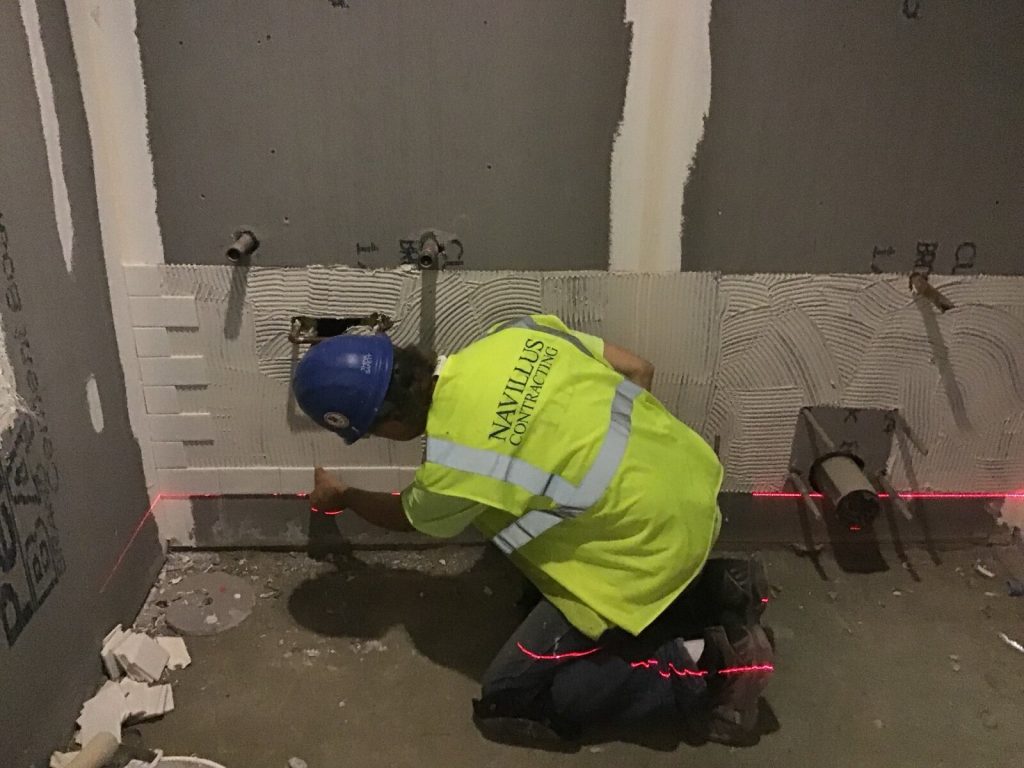 A tiler kneeling to set out ceramic wall tiles to a laser line over notched adhesive