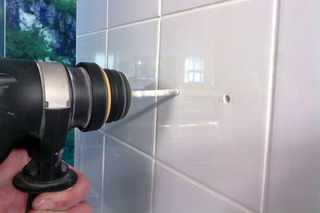 A drill with a tile bit held against a white tiled wall, next to an existing drilled hole