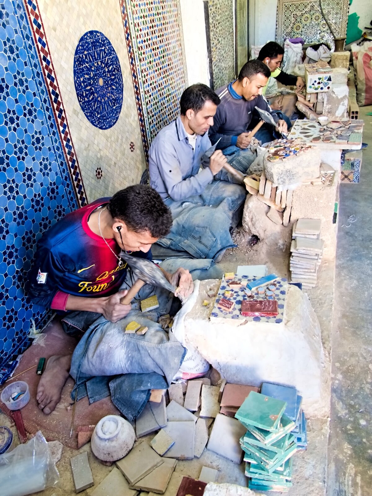 Craftsmen seated on the floor of a Moroccan workshop chipping glazed zellige tiles into shapes by hand