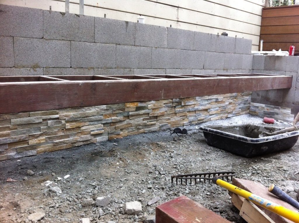 Split face stacked stone panels being fixed along the base of an outdoor block wall